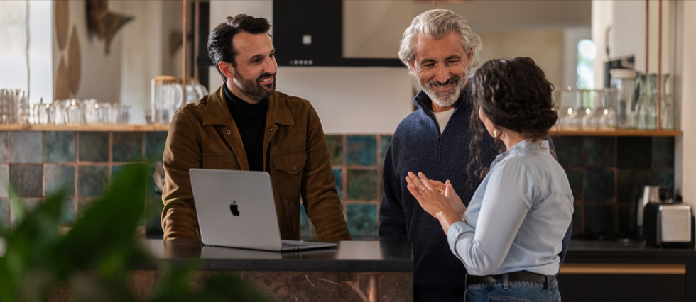 While a macbook sits on a table, a conversation occurs between two men and a woman.