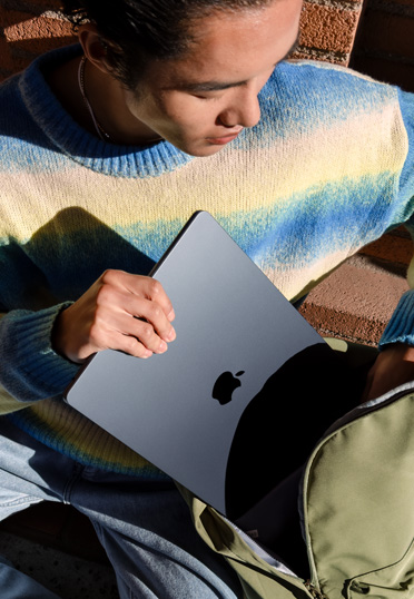 Un étudiant est assis à l’extérieur. Il sort un MacBook fermé de son sac à dos.